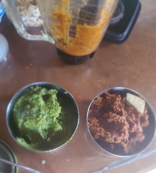 small stainless steel bowls on countertop containing green (pesto) mixture and burnt orange (sundried tomato) dips, a glass blender in background with orange colored remnants inside