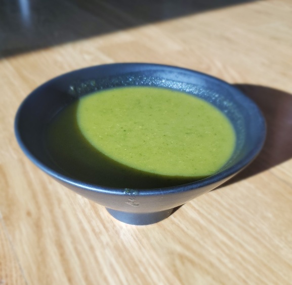 green puréed soup in black bowl on wooden counter, in a patch of sun